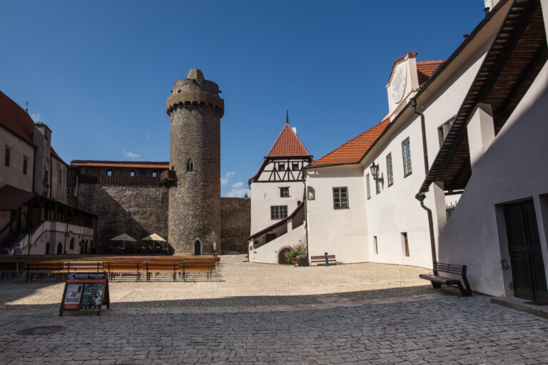 Historic stone tower and surrounding white buildings with red-tiled roofs in a cobblestone plaza; sunny day.