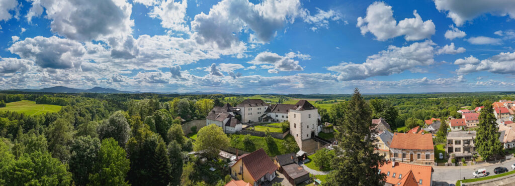 Panoramic aerial view of a rural village with white and red-roofed houses, green fields, and trees under a blue sky with clouds.