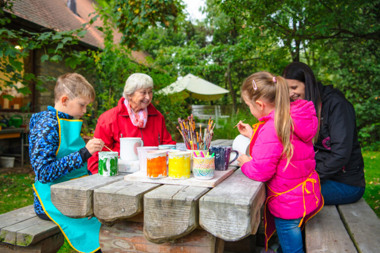 Children and two adults paint colorful pottery at a wooden picnic table in a leafy garden.