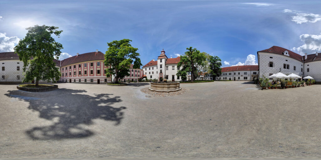 Central courtyard with a circular stone fountain, white and pastel buildings, and leafy trees beneath a bright blue sky.