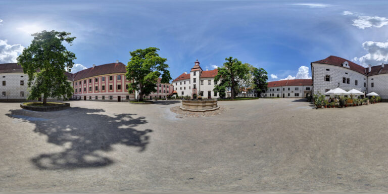 Central courtyard with a circular stone fountain, white and pastel buildings, and leafy trees beneath a bright blue sky.