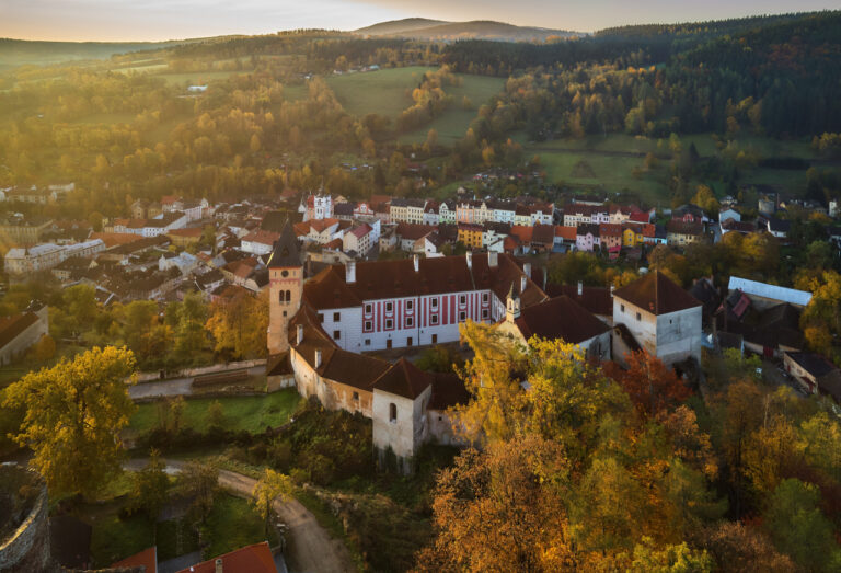 Aerial view of a historic town with a castle and red-tiled roofs, bathed in autumnal golden light.