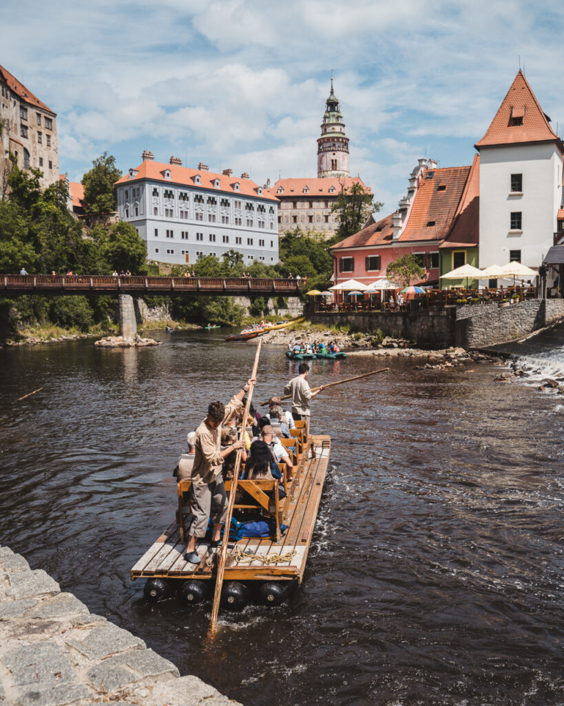 People ride a wooden raft with paddlers along a river in a historic town with red-tiled roofs and a bridge in the background.