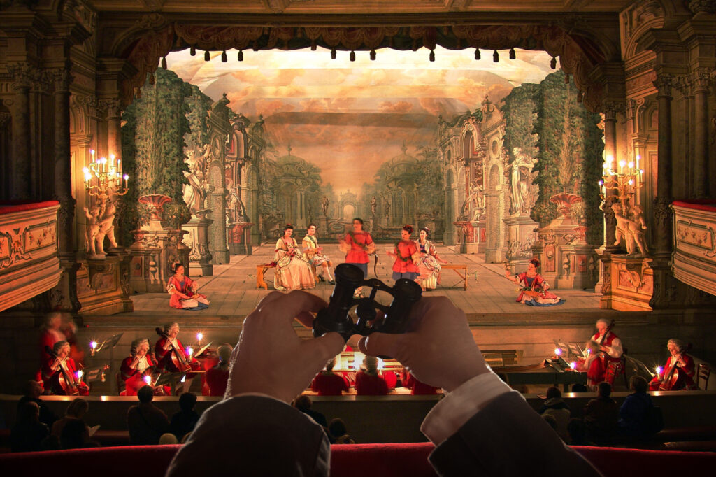 Audience watching a theatrical performance on an ornate stage; hands in the foreground hold binoculars to view the dancers and musicians.