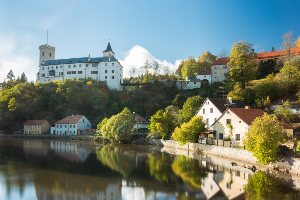 Castle on a hill overlooking a river, with white fortress, red-roofed houses, and autumn trees reflected in the water.