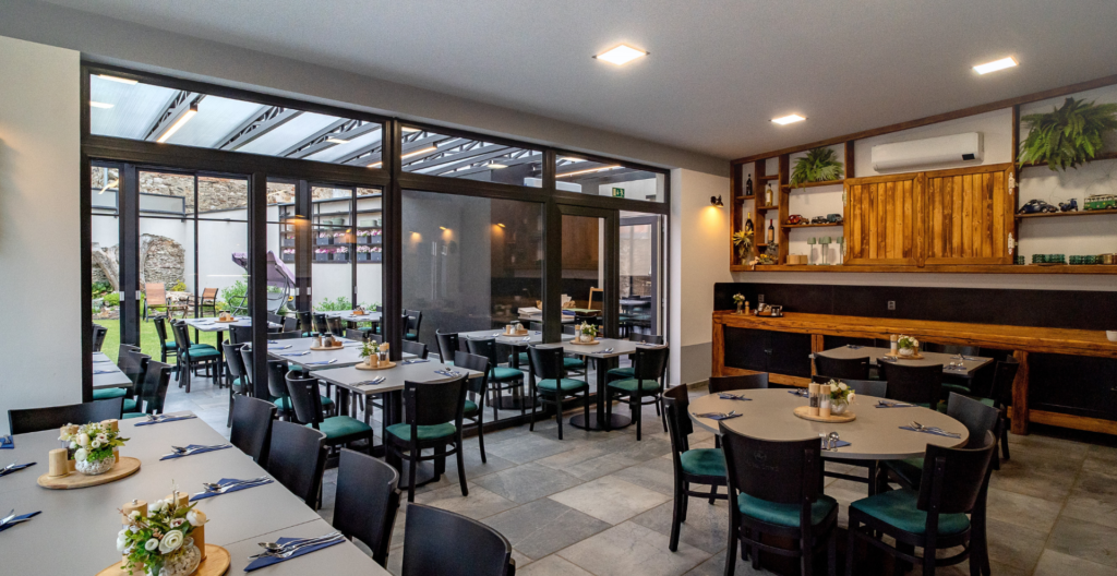 Restaurant dining area with grey tables, black chairs with green cushions, and a wooden bar along the right; glass walls reveal an outdoor patio, plants and seating beyond the glass panels.