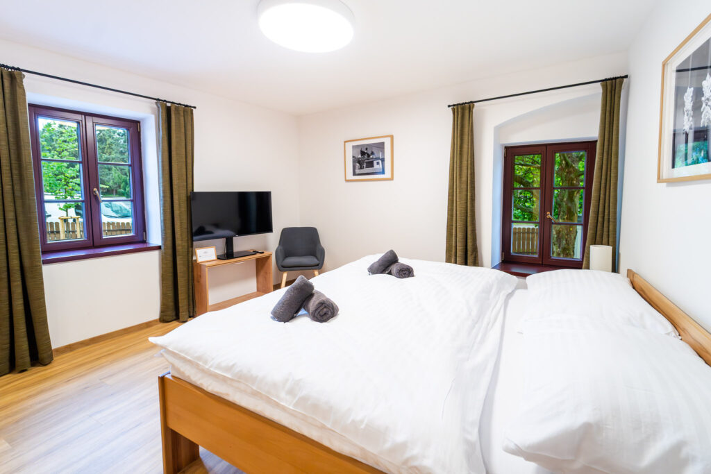 Twin beds in a bright hotel room with brown-framed windows, curtains, and a TV on a small desk
