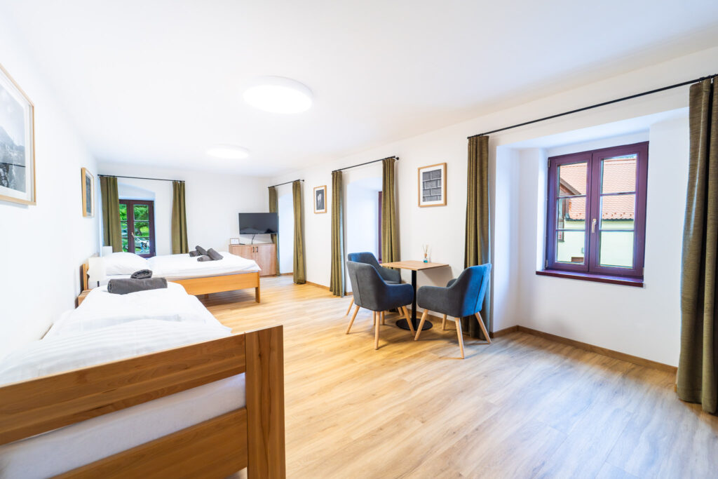 Bright hotel room with two single beds, wooden frames, white linens, and a seating area by a large window with green curtains.