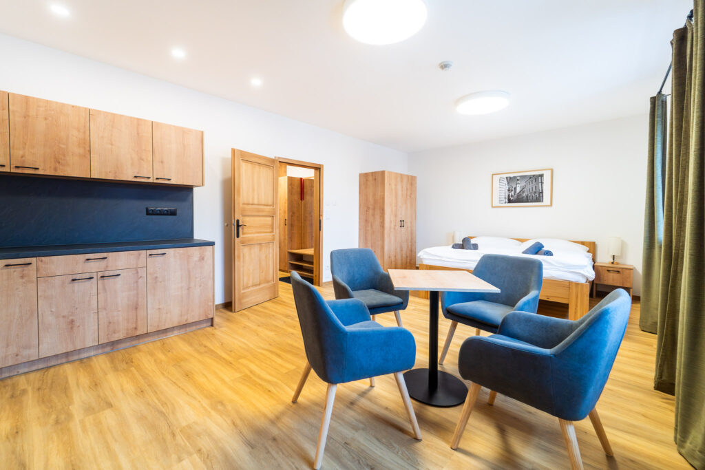 Hotel room with kitchenette on the left, a small wooden dining table and blue chairs, and a bed toward the back.
