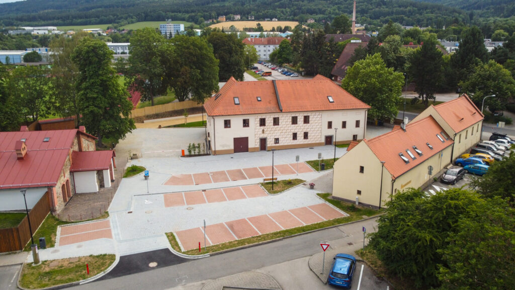 Aerial view of a town square with a large white building and red-tiled roofs, parked cars, and patterned brick paving.