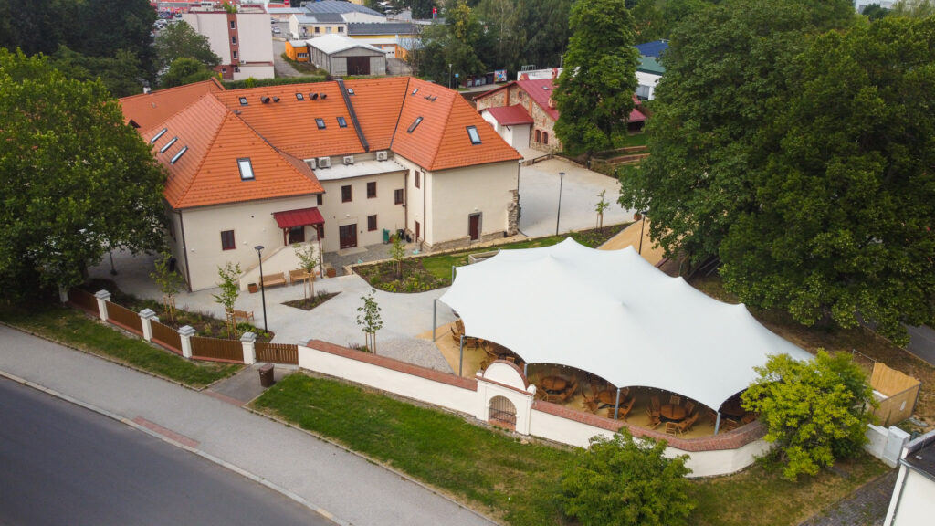 Aerial view of a beige building with red tile roofs, surrounding trees, and a large white event tent with wooden tables and chairs in a courtyard.