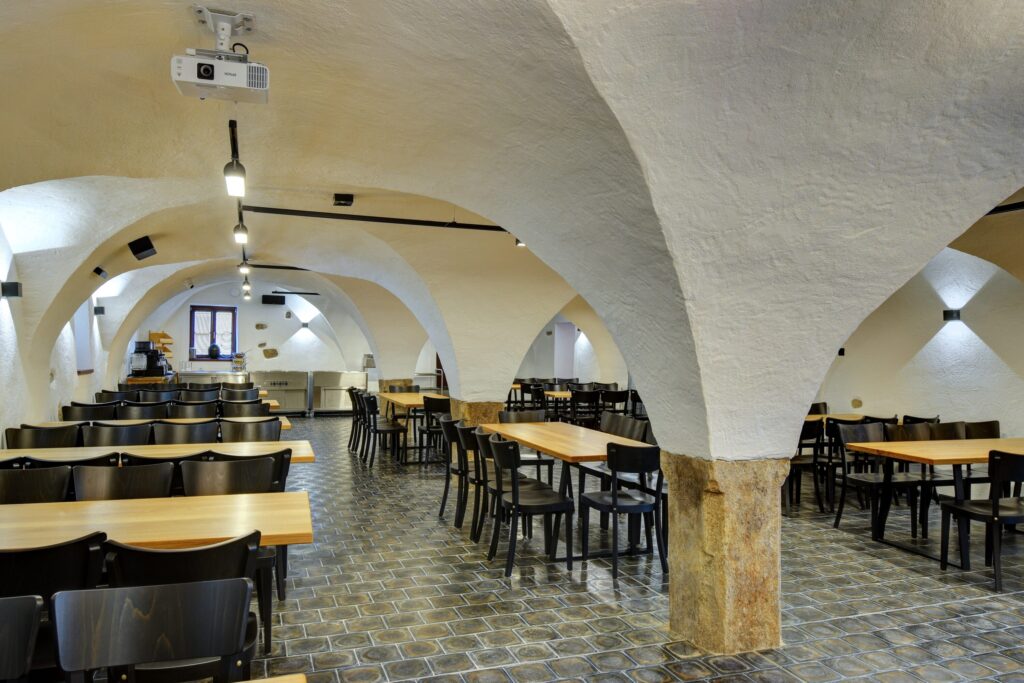 Interior of a vaulted dining hall with white arches, wooden tables and black chairs, and a projector on the ceiling.