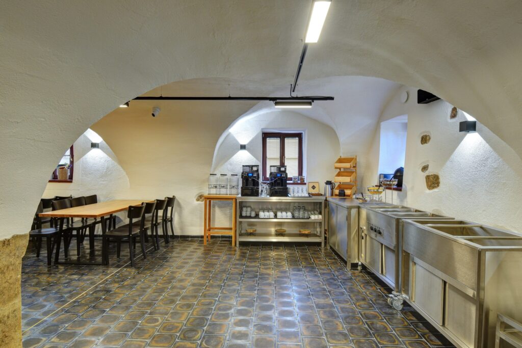 Cafè-style interior with arched white walls, wooden tables, black chairs, and a coffee station along the back wall.