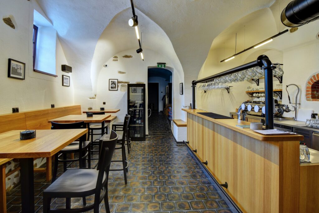 Café interior with arched white ceiling, wooden tables and black chairs, and a bar with glassware on the right.