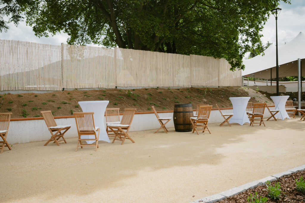 Outdoor event setup with wooden chairs around white cocktail tables on a sandy patio, a bamboo fence behind, and trees overhead.