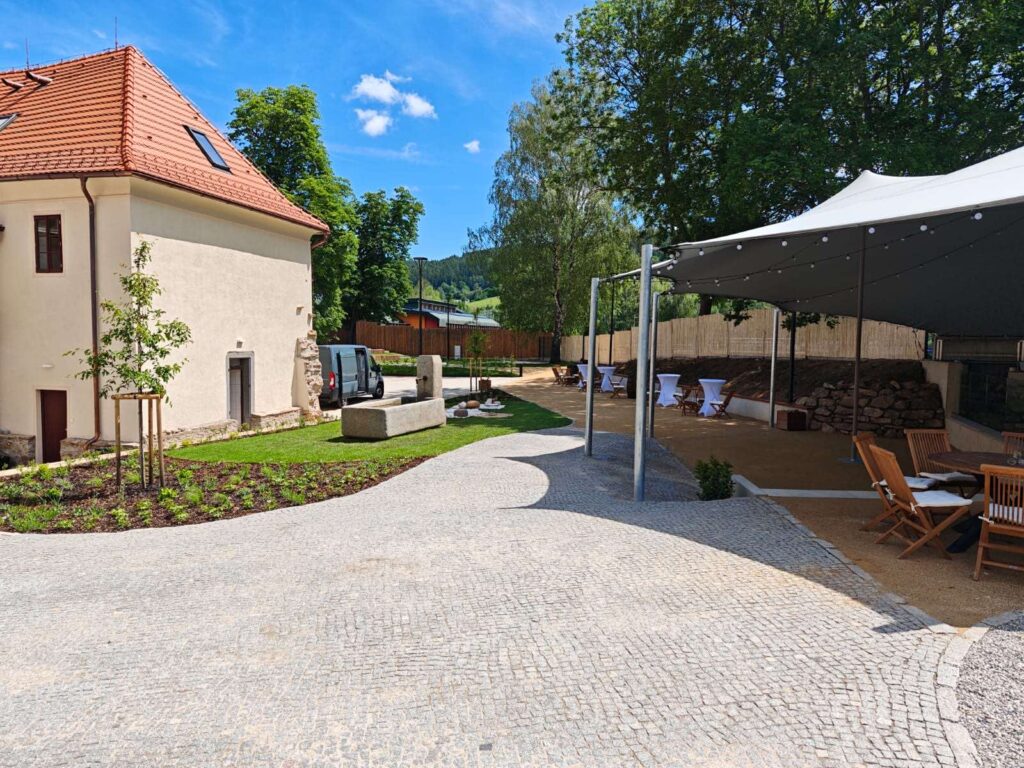 Courtyard scene with a cream building and red-tiled roof on the left, a cobblestone path, garden beds, and a white canopy with string lights on the right, plus wooden chairs and tables set up for an event. A van is parked near the building, and green hills are in the distance under a blue sky.