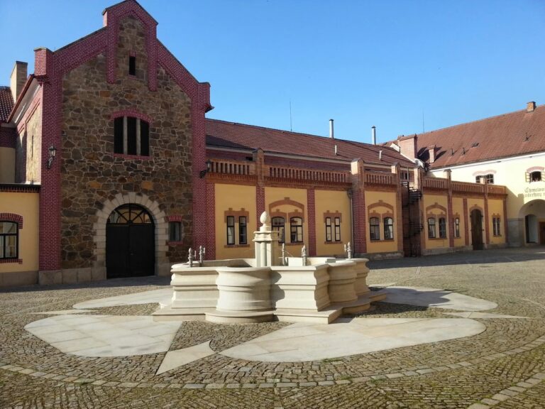 Historic courtyard with a central ornate stone fountain, surrounded by yellow-walled buildings with red brick trim and a blue sky above.